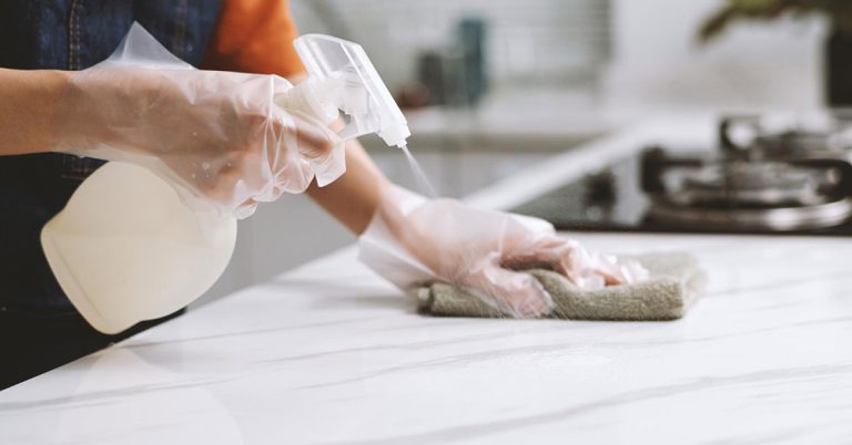 Person demonstrating how to clean natural stone countertops using pH-neutral stone cleaner spray and microfiber cloth with proper protective gloves in modern kitchen